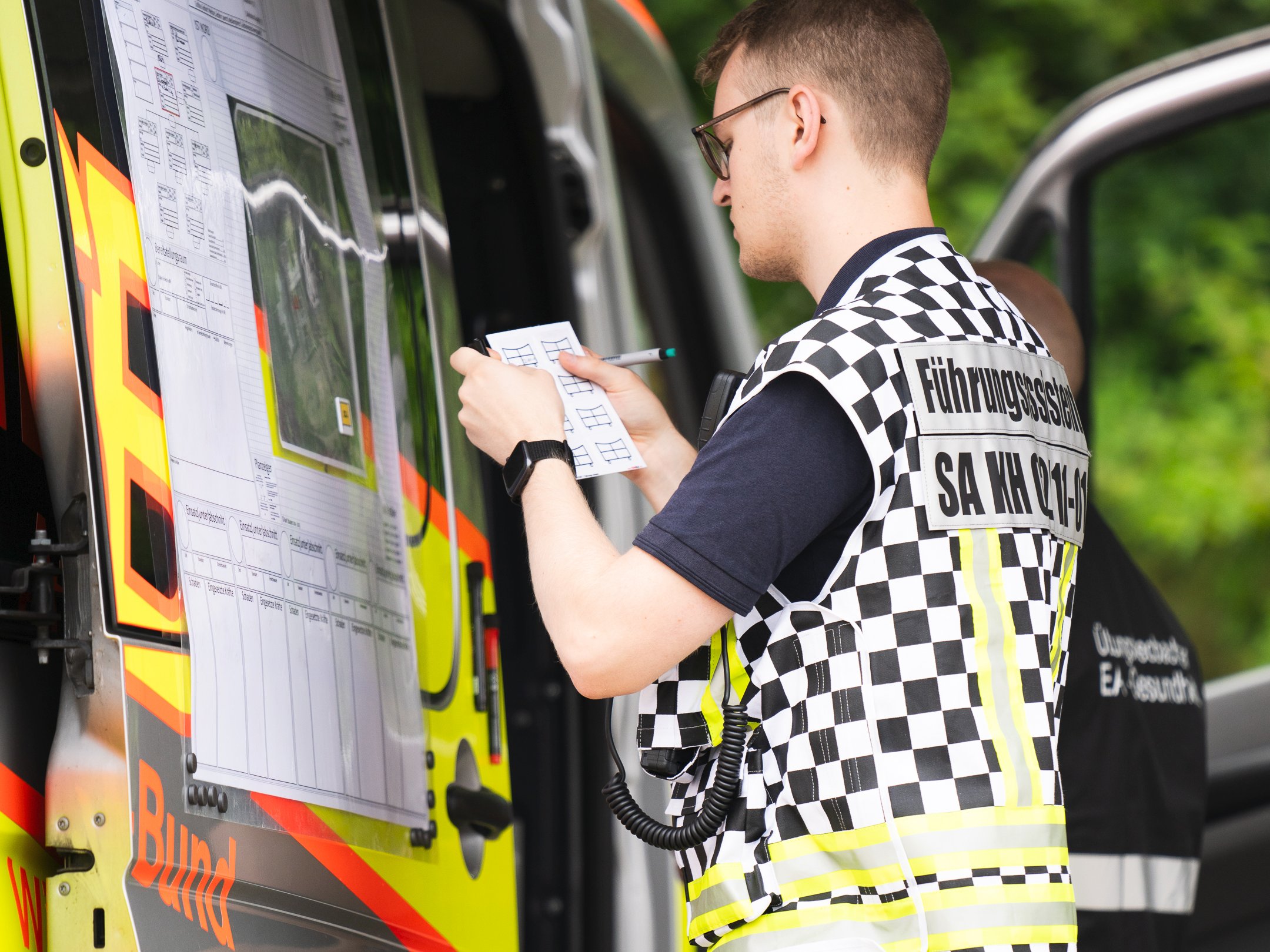 Ein Führungsassistent in schwarz-weiß-karierter Weste steht mit Zettel und Stift an der Lagekarte vor dem Einsatzleitwagen