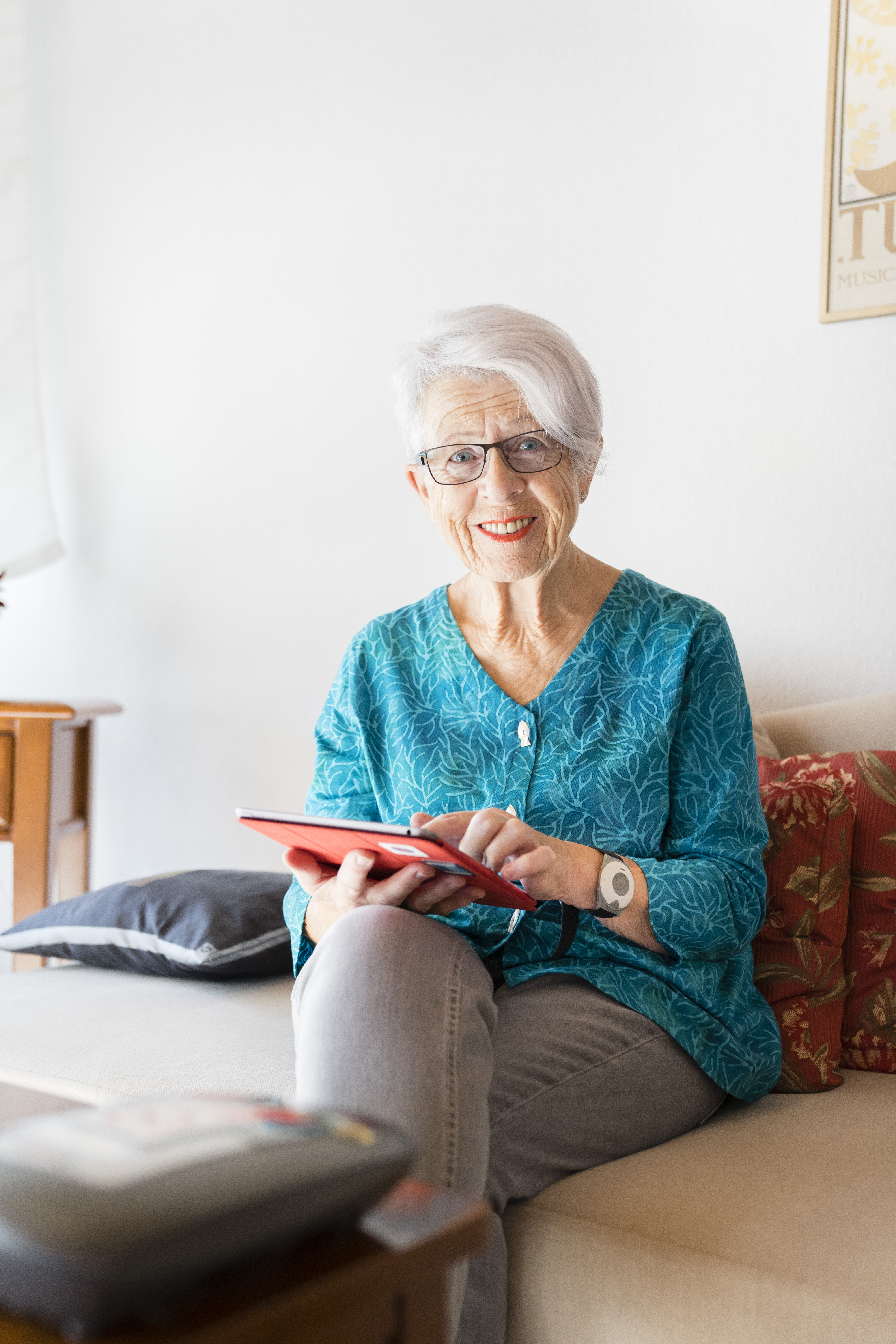 Ältere Dame sitzt auf einem Sofa. Sie hat ein Tablet in der Hand und einen Hausnotrufsender am Handgelenk.