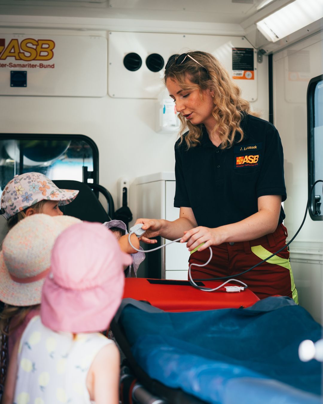 Eine Notfallsanitäterin steht mit 3 Kindern im Rettungswagen. Sie hat einen Sauerstoffclip in der Hand und erklärt etwas.